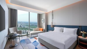 Quarto de hotel moderno com uma cama grande, área de estar com vista e paisagem urbana visível pela janela.