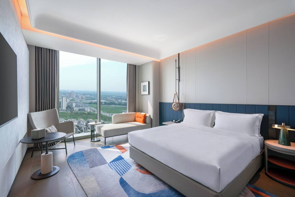 Una habitación de hotel moderna con una cama grande, un área de descanso con vistas y un paisaje urbano visible a través de la ventana.