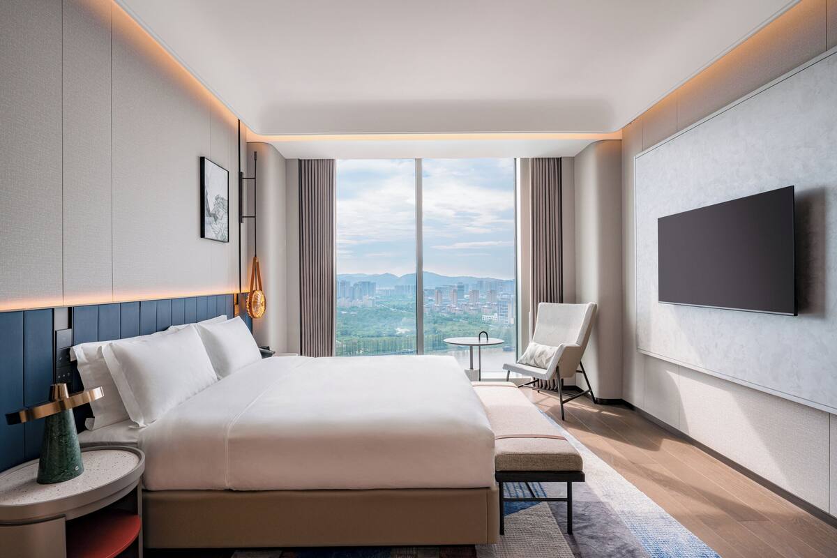 Una habitación de hotel moderna con una cama grande, televisión, una silla y vistas de la ciudad.