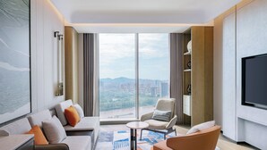 Quarto de hotel moderno com uma janela grande, um sofá, uma cadeira, uma mesa pequena e uma televisão de tela plana.