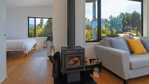 Un salon moderne avec un poêle à bois, un canapé gris agrémenté de coussins, un lit dans la pièce adjacente et de grandes fenêtres offrant une vue sur la verdure.