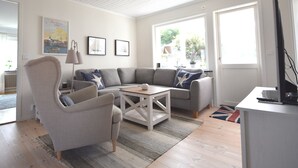 Un salón con un sofá gris, una mesa de centro blanca y un sillón gris. Hay cuadros enmarcados en la pared y una ventana con vistas a zonas verdes.