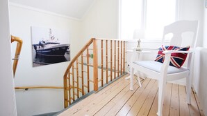 Una escalera con barandilla de madera, una silla blanca con cojín con la bandera del Reino Unido, y un cuadro de un barco en la pared.