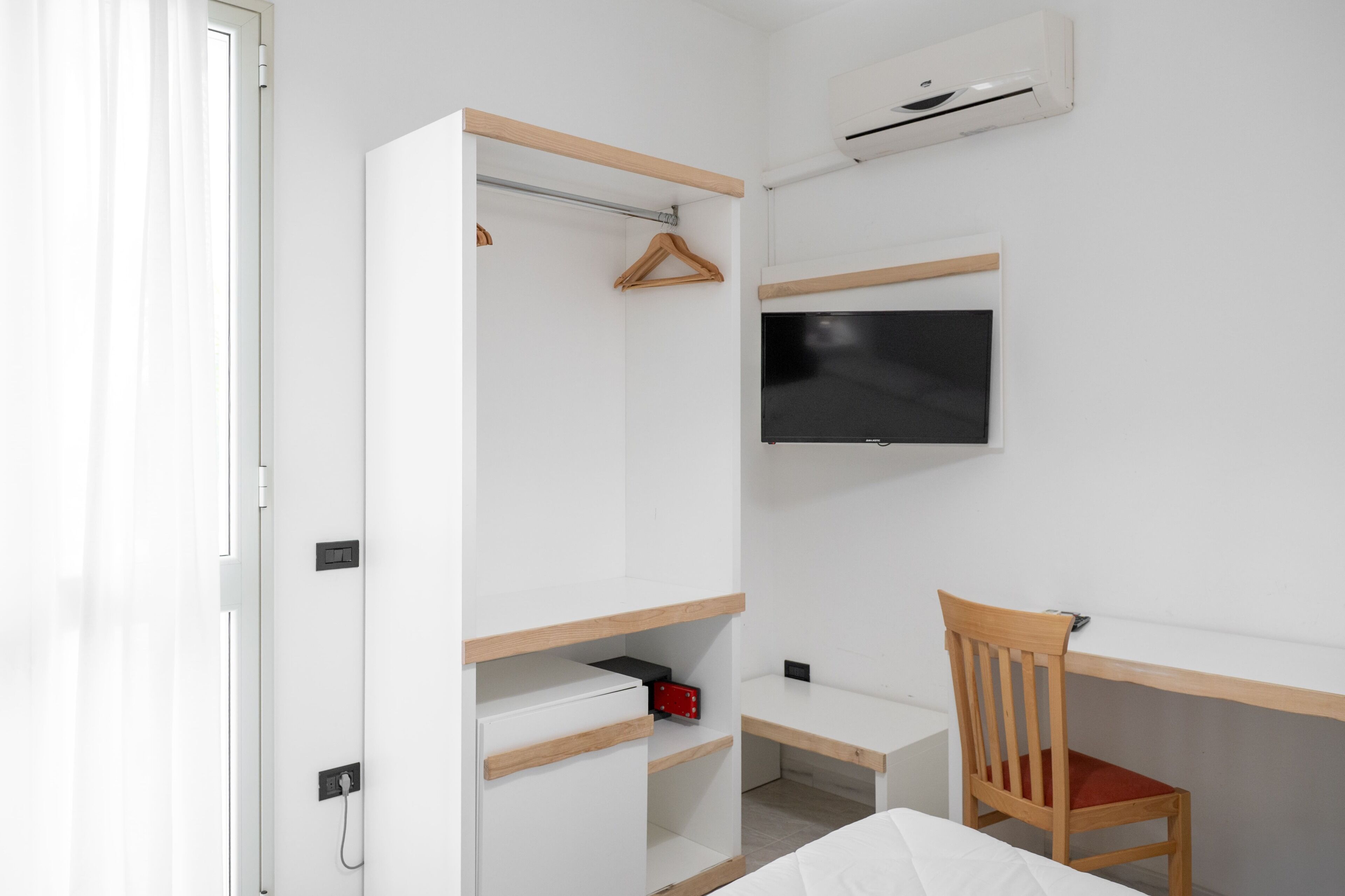 A bedroom with a bed, a desk, a chair, a wardrobe, a TV, and an air conditioning unit.
