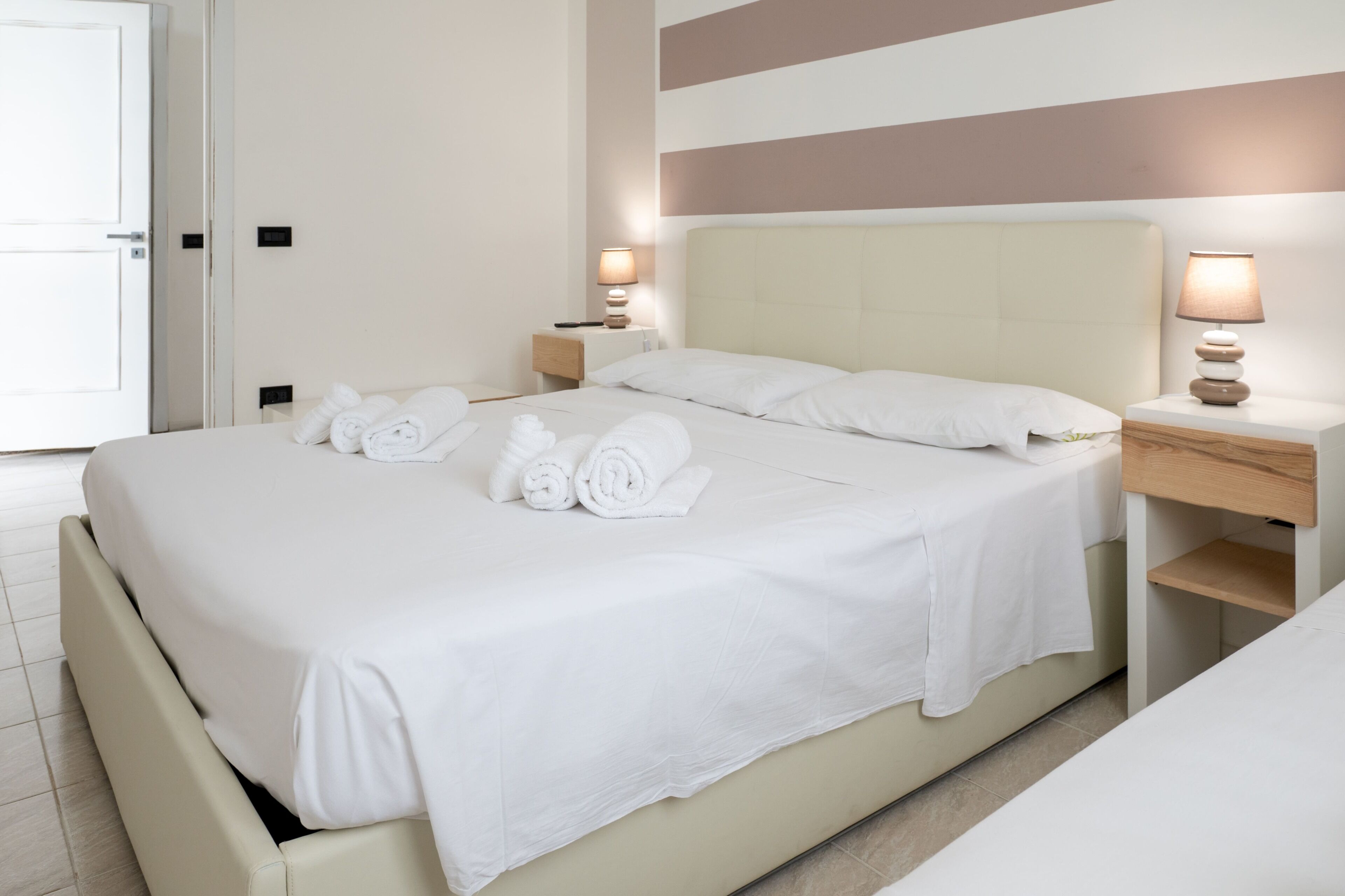 A neatly made bed with white linens and rolled towels, a bedside table with a lamp, and a door to the left.