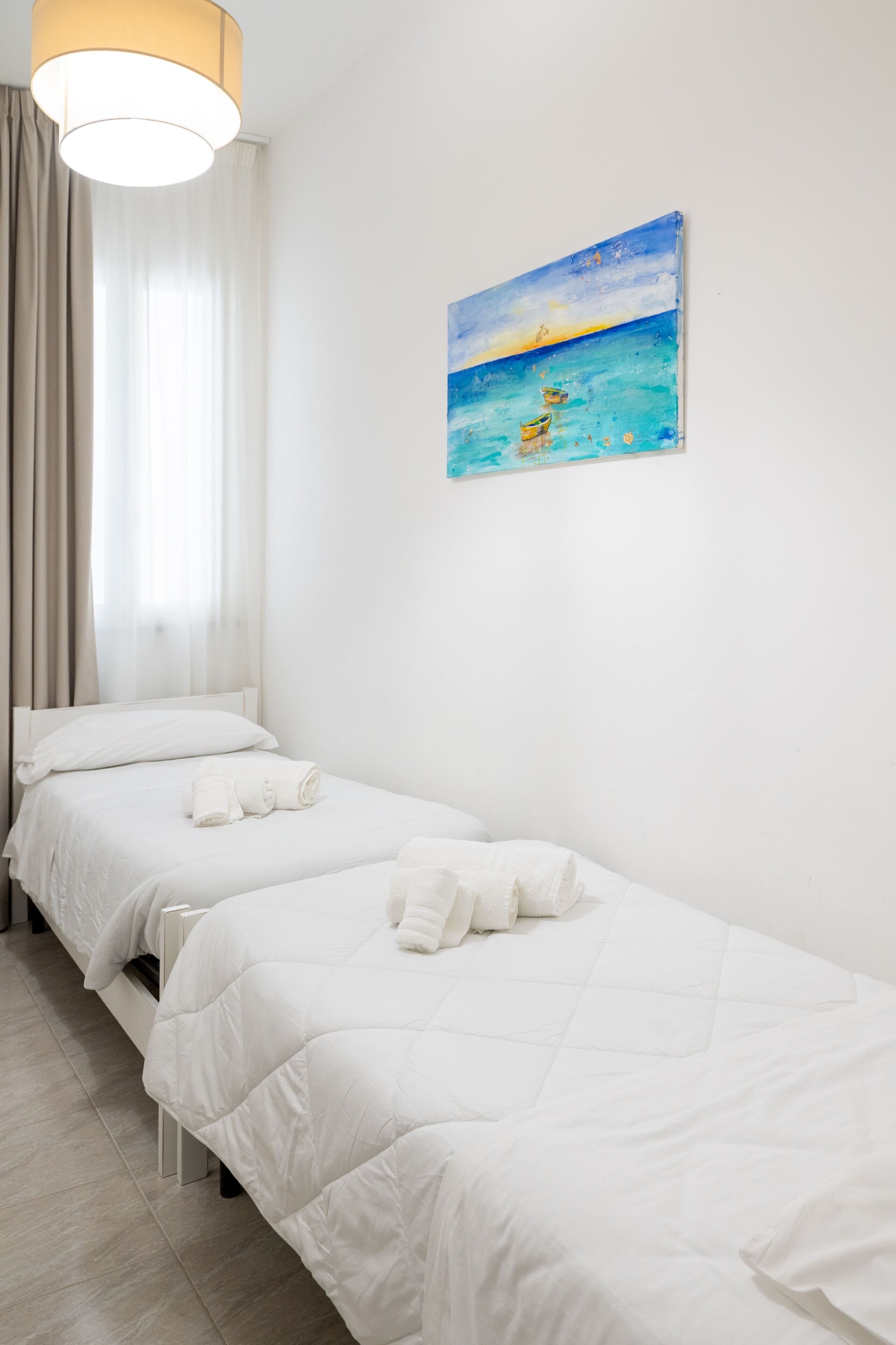 A hotel room with two single beds, a painting of a beach scene, a window with curtains, and a light fixture.