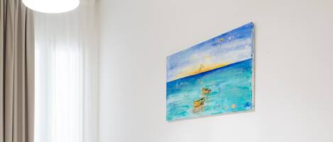 A hotel room with two single beds, a painting of a beach scene, a window with curtains, and a light fixture.