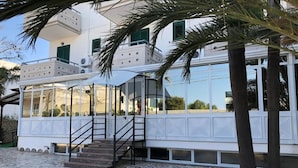A building with a white exterior, a covered outdoor area with a staircase, and palm trees. - Hotiday Porto Cesareo (Porto Cesareo)