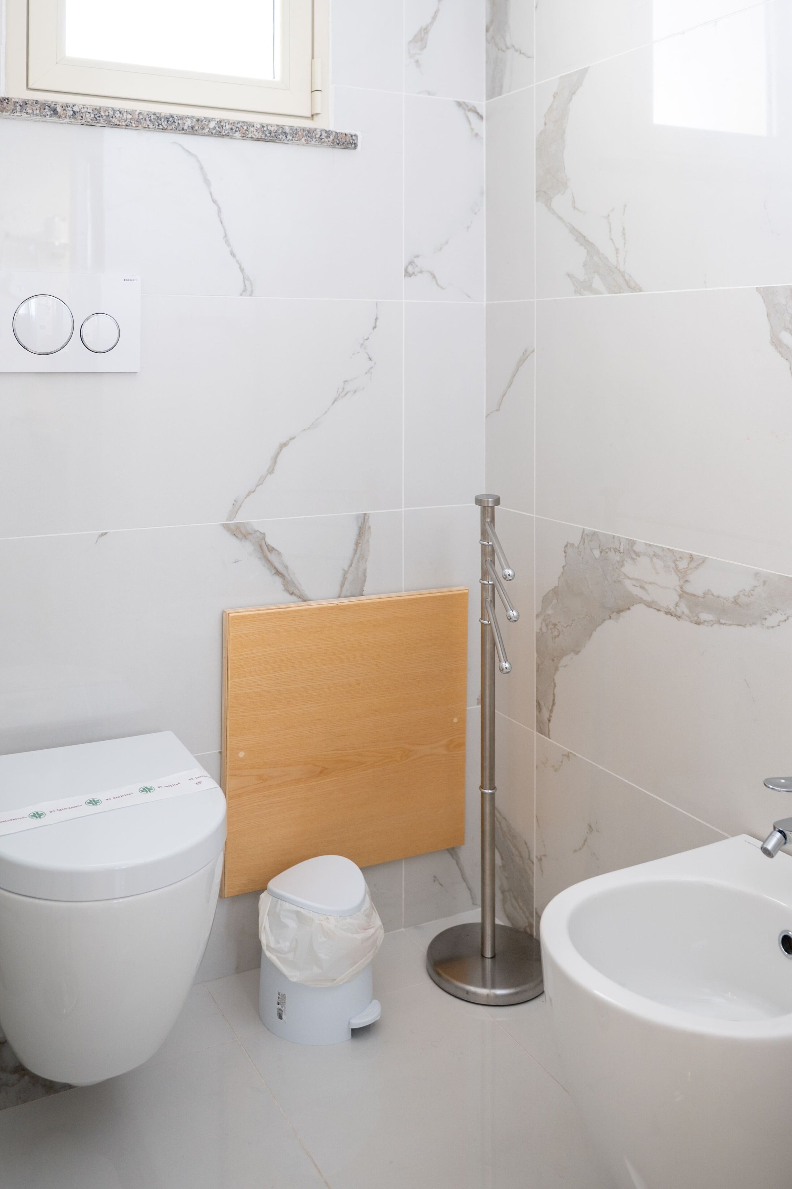Un baño con inodoro, bidé y lavamanos, paredes de mármol y un panel de madera.