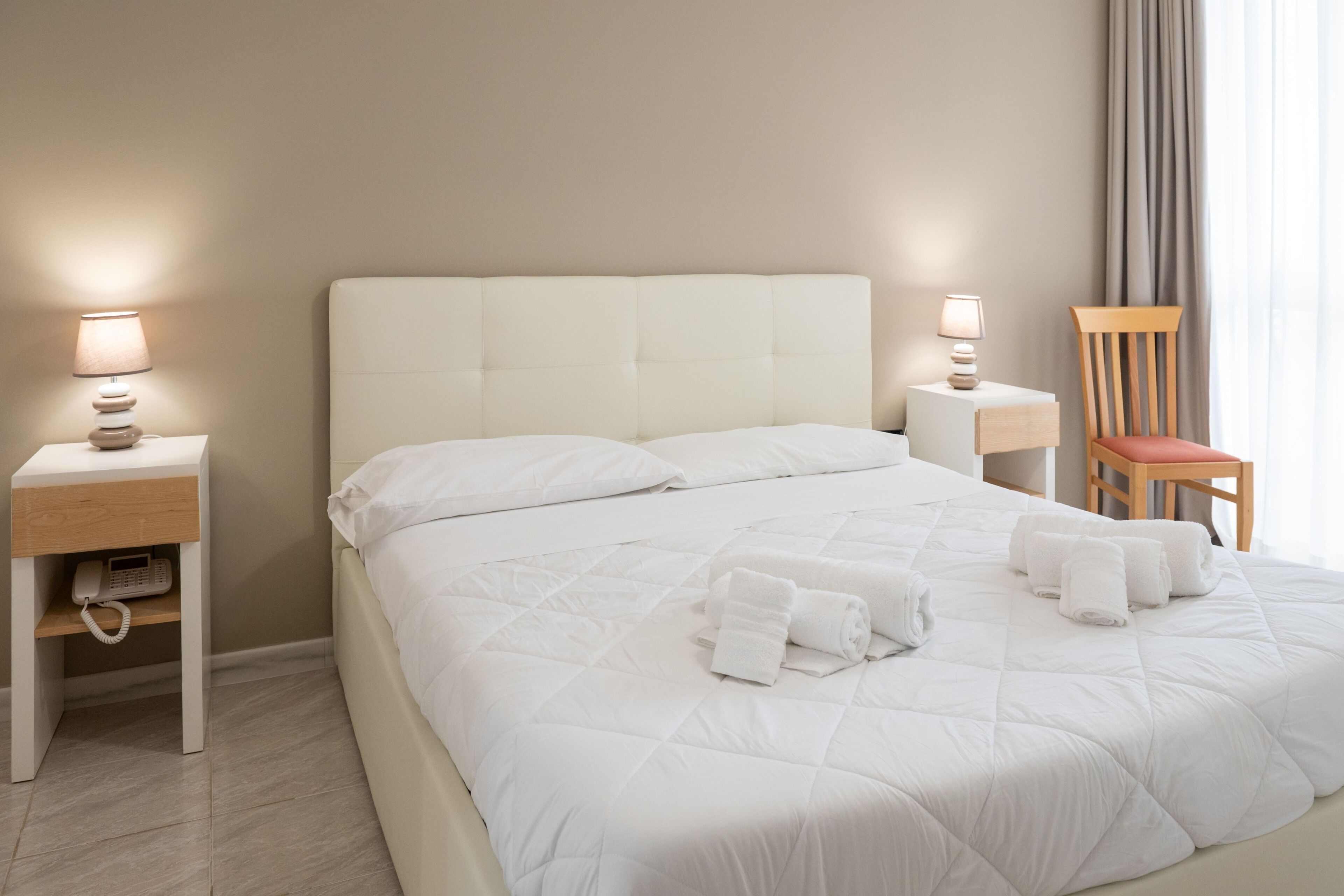 Una cama bien hecha con ropa blanca, dos mesitas de noche con lámparas, una silla y un teléfono al lado izquierdo.