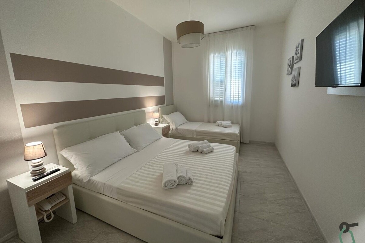 A hotel room with a large bed, a smaller bed, a bedside table with a lamp, and a TV mounted on the wall.