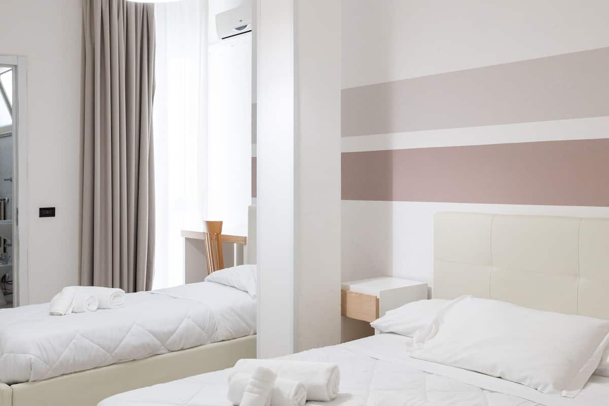 A hotel room with two beds, a hanging light fixture, a bedside table, and a view of the outside.