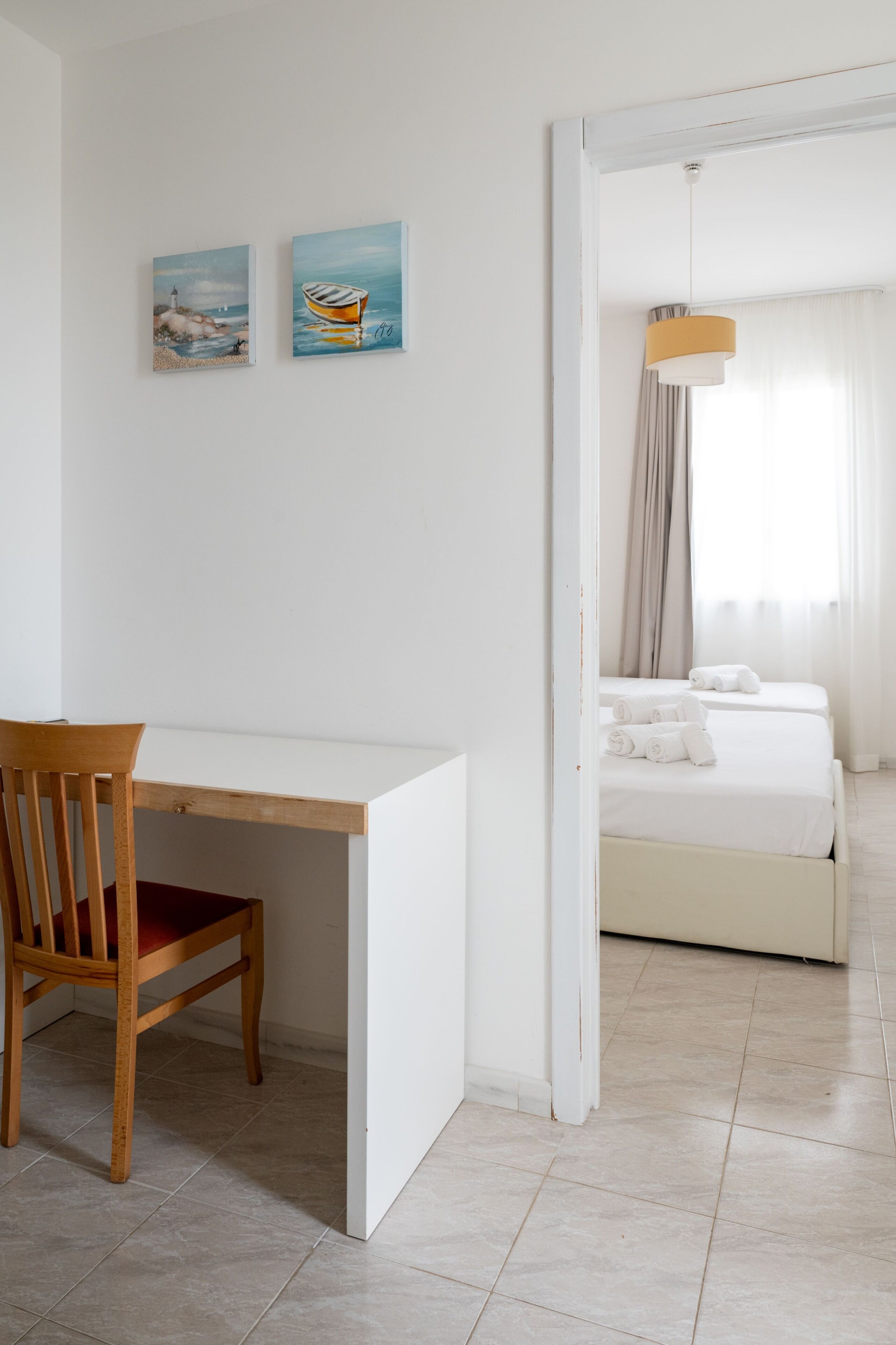 A room with a wooden desk, a chair, a bed with white linens, and two paintings on the wall.
