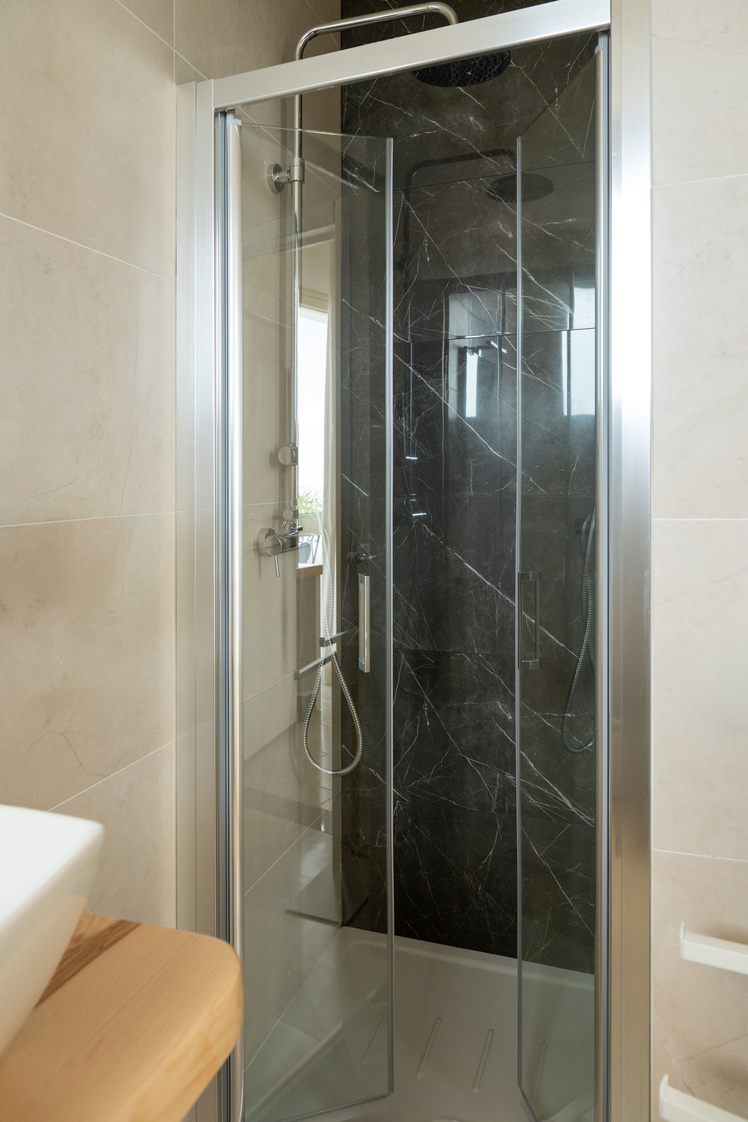 A glass-enclosed shower with a marble interior, a white sink, and a wooden bench.