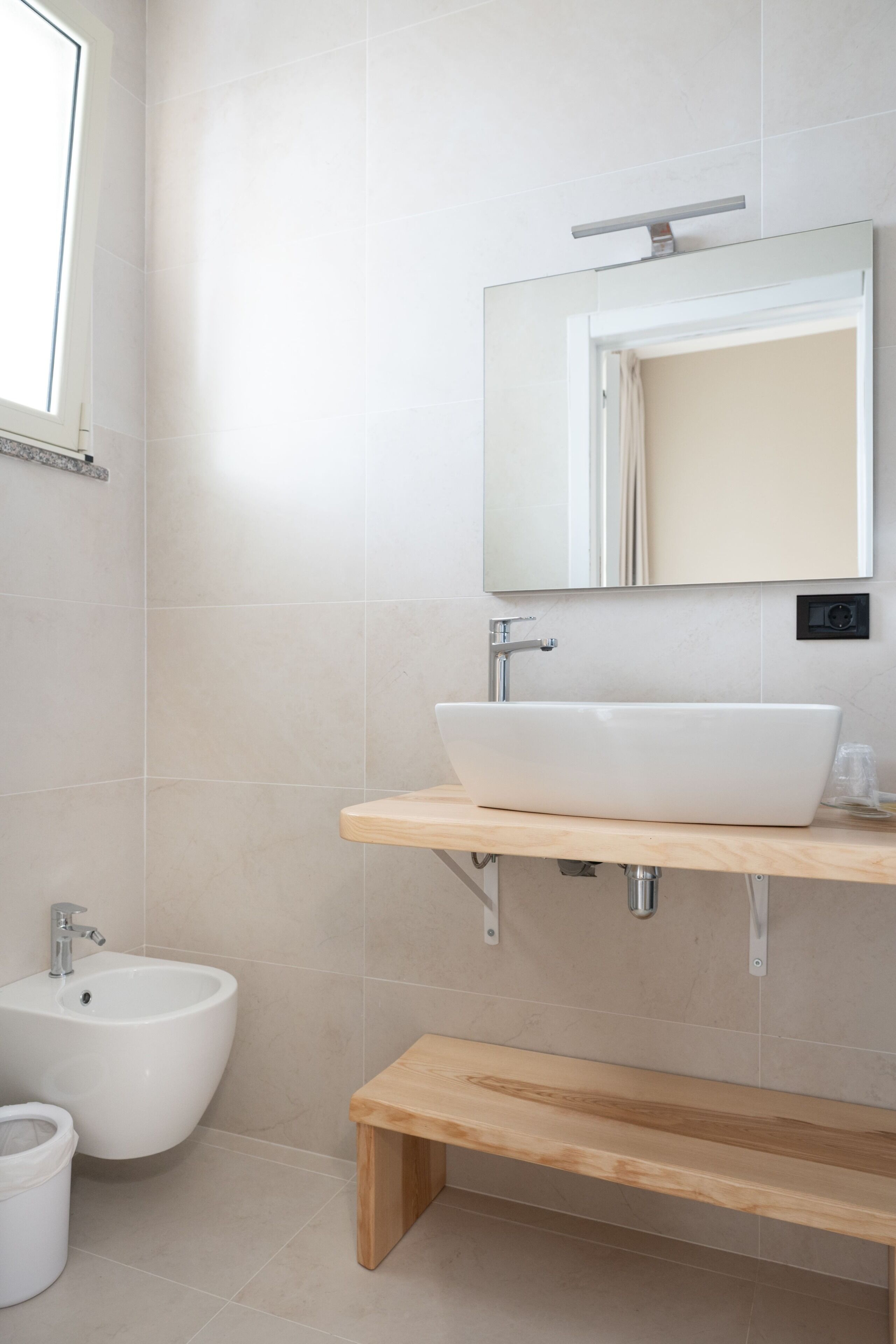 Un baño moderno con un lavamanos blanco, un bidé, un banco de madera y un espejo en la pared.