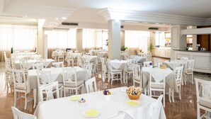 A spacious dining area with multiple tables and chairs, a kitchen in the background, and large windows allowing natural light. - Hotiday Porto Cesareo (Porto Cesareo)