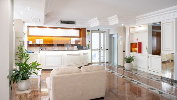 A hotel lobby with a reception desk, a sofa, a large potted plant, and a marble floor. - Hotiday Porto Cesareo (Porto Cesareo)