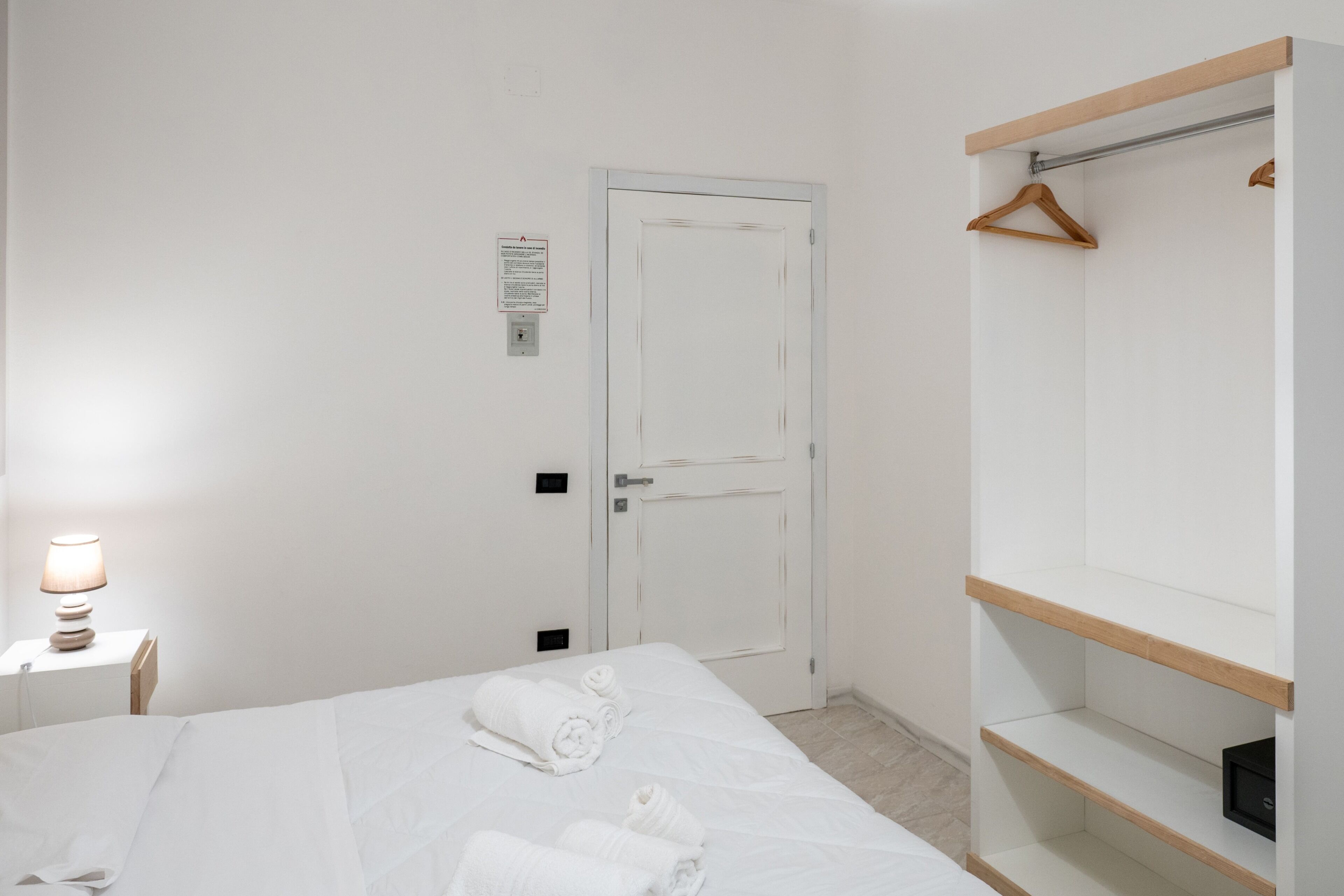 A hotel room with a bed, a bedside table with a lamp, a wardrobe with hanging space, and a door with a sign.