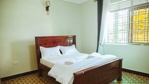 Cama bem arrumada em quarto com piso de madeira estampado, luminária fixada na parede e janela com grades.
