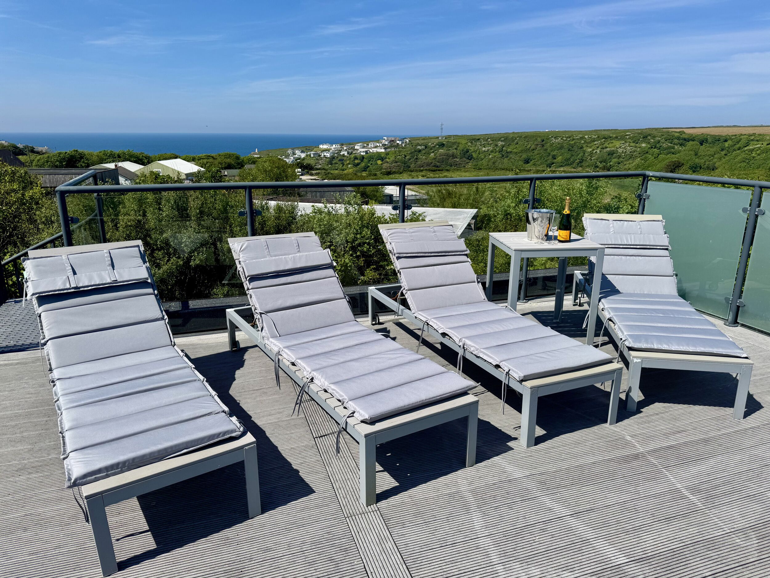 Eine Dachterrasse mit Liegestühlen, einem Tisch mit einer Flasche und Blick auf Meer und Grünfläche.