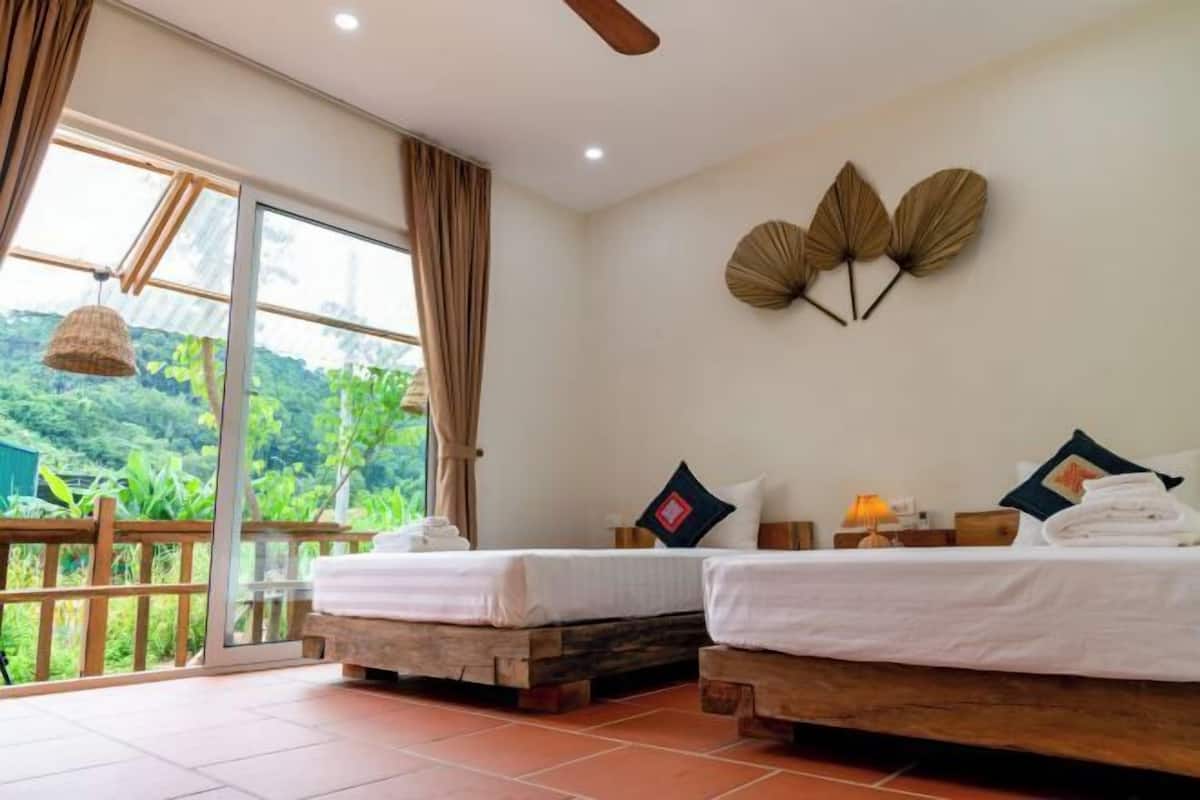 A room with two beds, a large window with curtains, a ceiling fan, and a view of greenery outside.