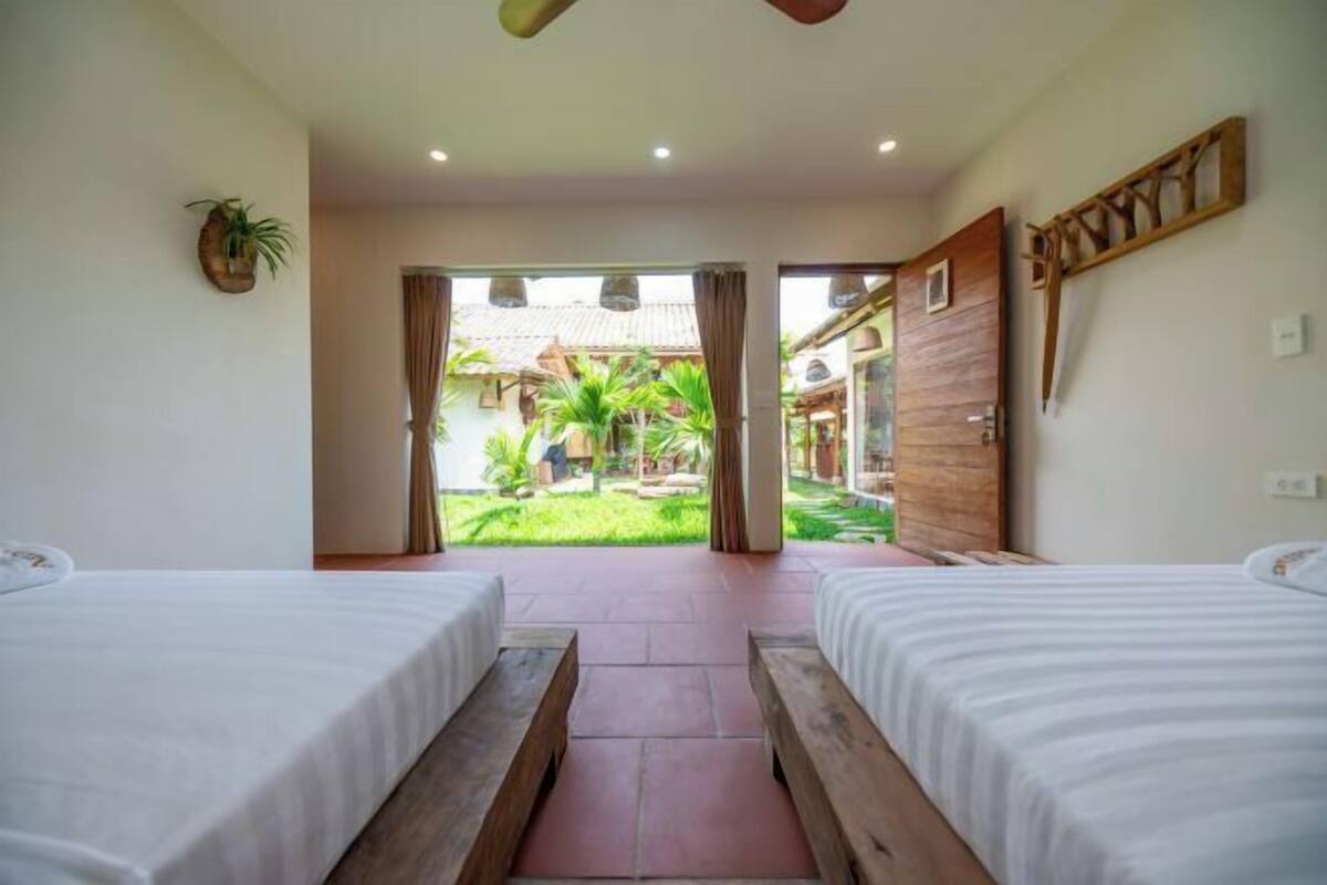 A room with two beds, a wooden bench, and a view of a garden and building outside.