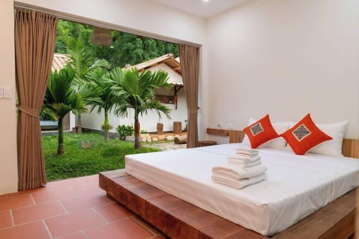 A bedroom with a bed, two pillows, a nightstand, and a view of a garden through an open door.