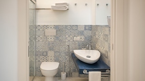 Een badkamer met een wit toilet, een wand betegeld met blauw-wit patroon, een witte wastafel met een bijzondere vorm, en een handdoekrek met opgevouwen handdoeken.