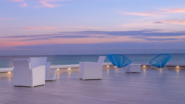 Een terras op het dak met witte rieten stoelen en een blauwe stoel, met uitzicht op de zee.