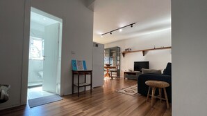 A spacious living area with a wooden floor, a sofa, a television, and a dining table with chairs.