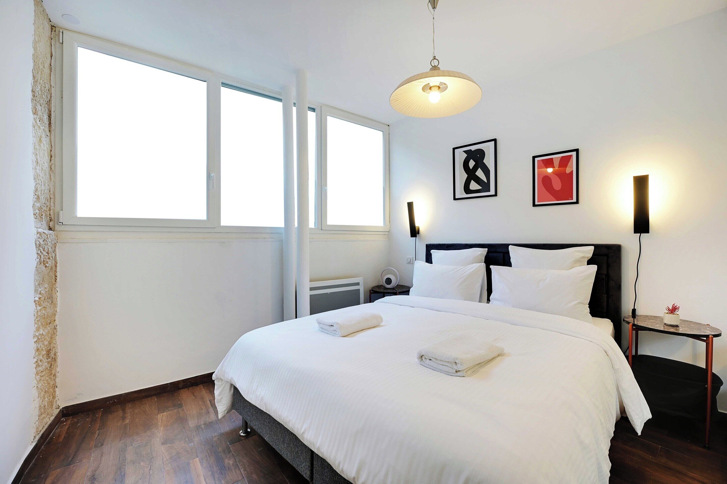 A neatly made bed with white linens, a bedside table with a lamp, and a wall-mounted artwork.