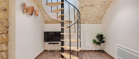Un escalier en colimaçon avec des marches en bois et des rampes en métal, dans une pièce aux murs en pierre, équipée d’un téléviseur à écran plat et agrémentée de plantes en pot.