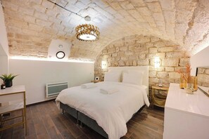 A bedroom with a stone ceiling, a bed with white linens, a bedside table, a small plant, and a radiator. - Charming Apartment - 2bd/6p - Port Royal (Paris)