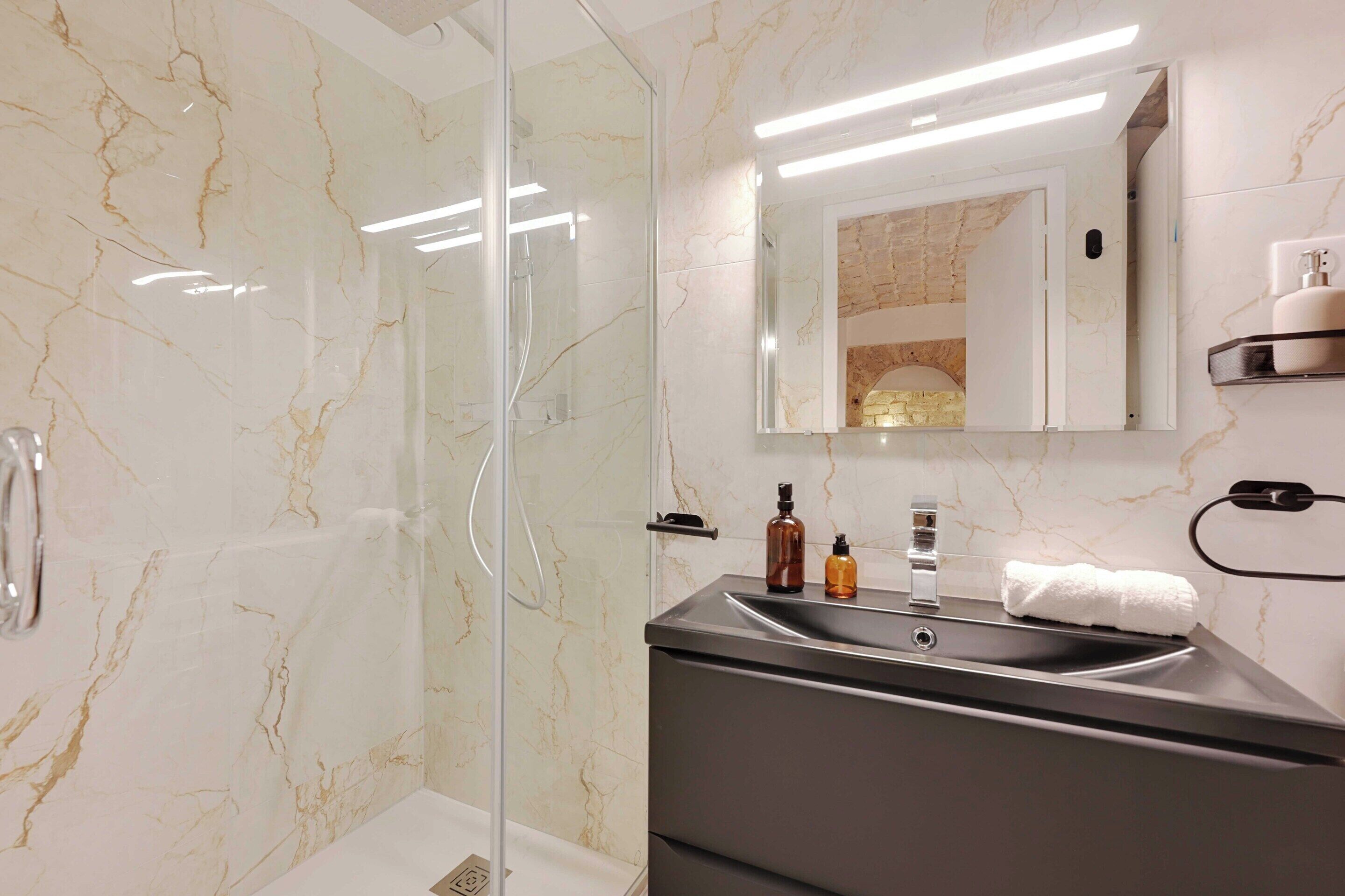 A modern bathroom with a glass-enclosed shower, a marble wall, a sink with a mirror, and toiletries on the countertop.