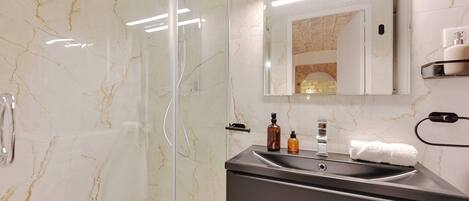 A modern bathroom with a glass-enclosed shower, a marble wall, a sink with a mirror, and toiletries on the countertop.