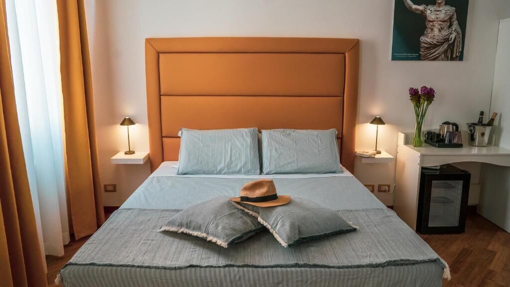 A neatly made bed with a brown headboard, two bedside lamps, and a hat on the pillows.