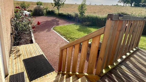 A wooden deck with a bench, a paved path, and a view of a field and trees.
