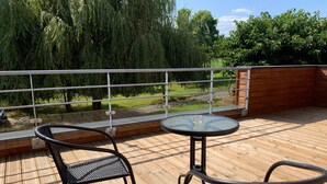 A wooden deck with a glass table and two wicker chairs, overlooking a grassy area and trees.