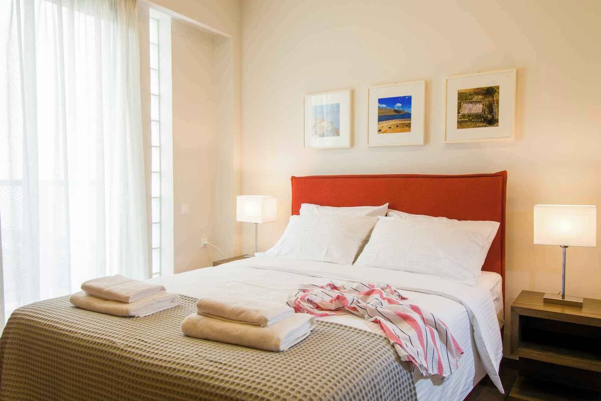 A hotel room with a bed, two bedside tables, a window with curtains, and three framed pictures on the wall.