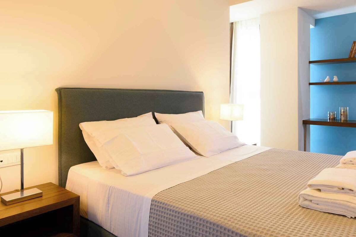 A neatly made bed with white linens and pillows, a bedside table with a lamp, and a small shelf with toiletries.