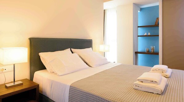 A neatly made bed with white linens and pillows, a bedside table with a lamp, and a small shelf with toiletries. - Elounda Colour Apartments (Elounda)