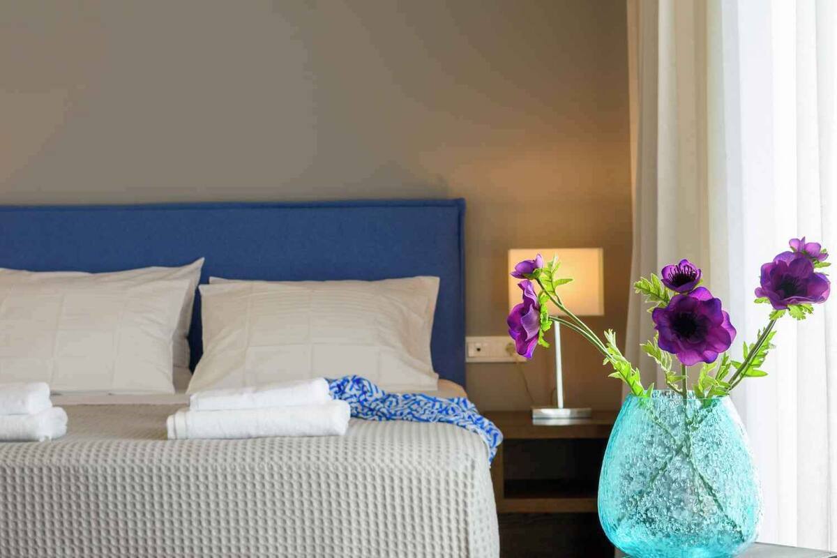 A neatly made bed with white linens and two pillows, a blue headboard, and a vase with purple flowers on a bedside table.