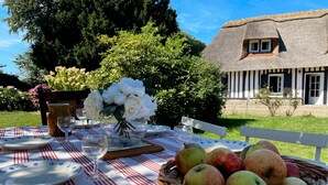 A picnic setup with a table set for two, featuring a striped tablecloth, plates, wine glasses, a basket of apples, and a vase of flowers. - Le Clos de Leger, a charming hamlet (Le Mesnil-Durdent)