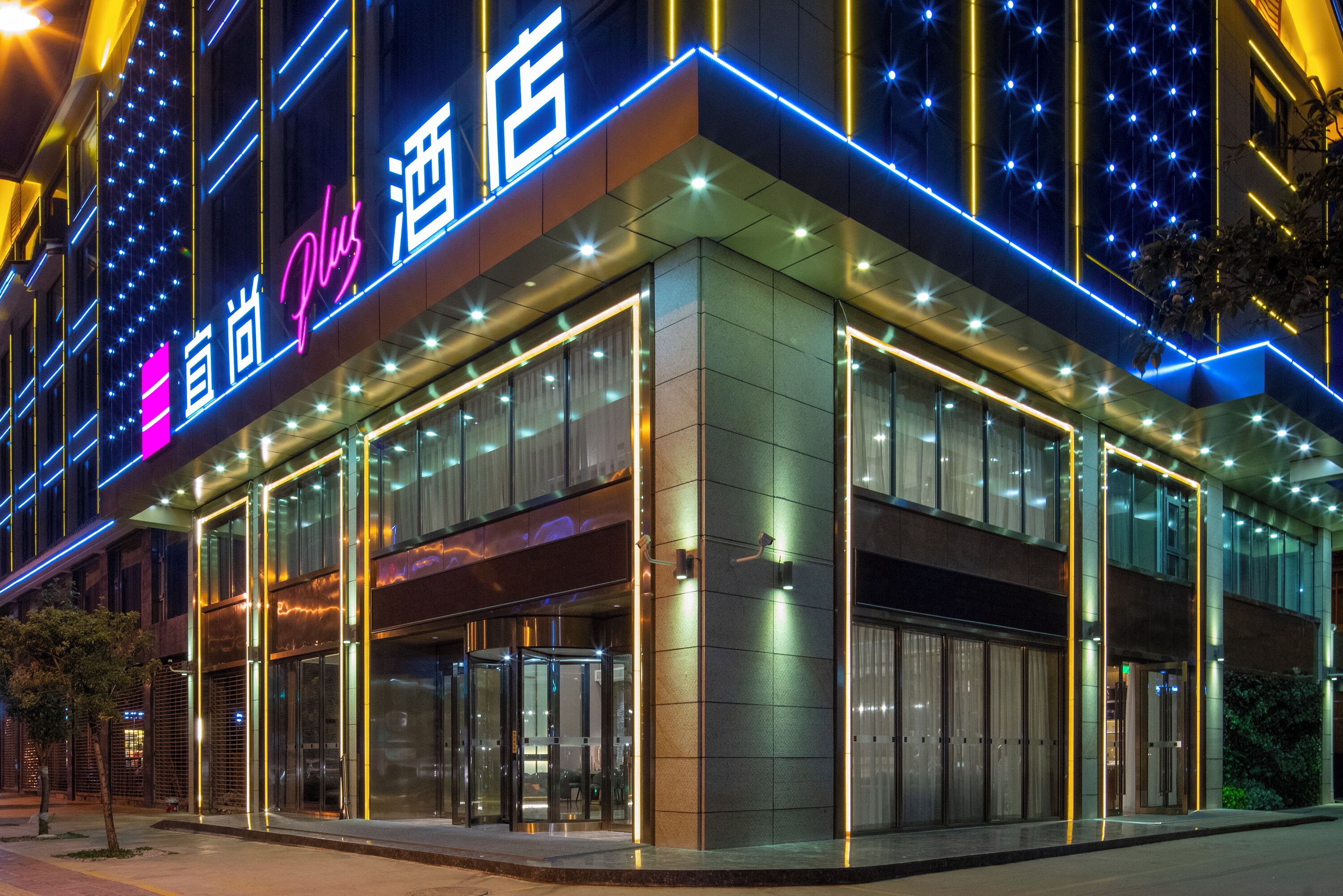 Un hôtel moderne doté d’une façade en verre, d’une enseigne lumineuse et d’une porte tournante à l’entrée.