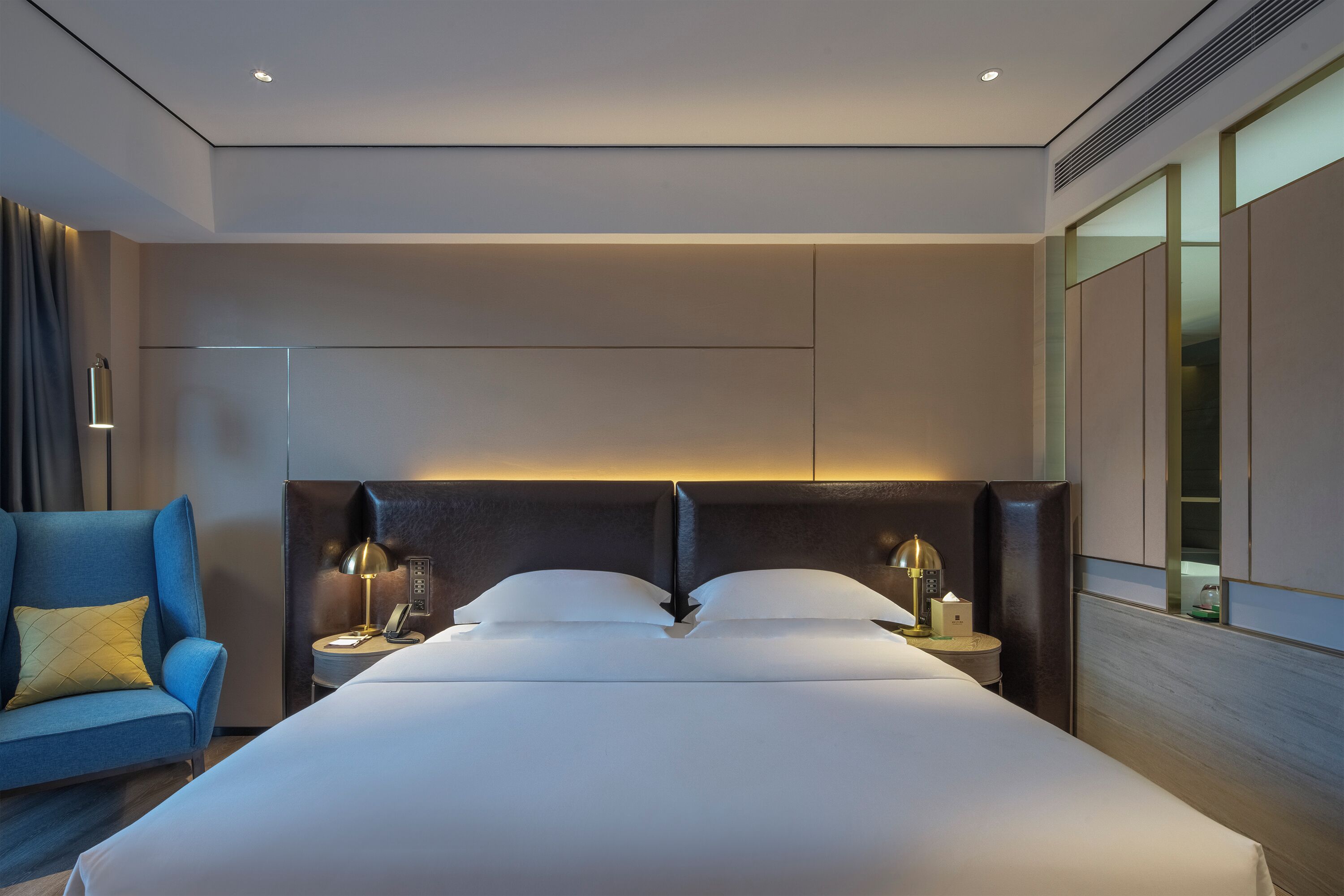 Une chambre d’hôtel moderne avec un grand lit, une table de chevet avec une lampe, un fauteuil bleu et une vue sur le paysage urbain par la fenêtre.