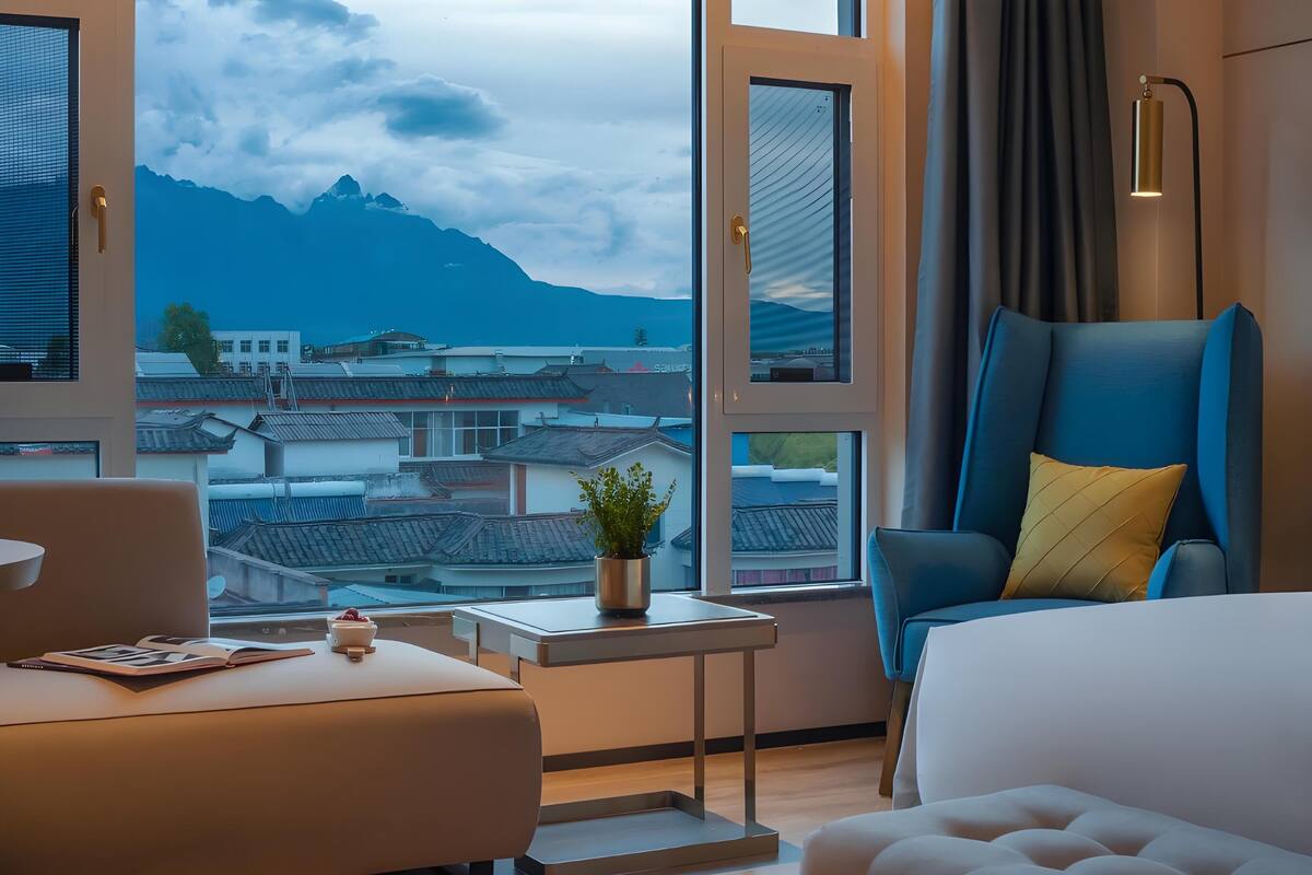 Habitación de hotel moderna con un gran ventanal, un sillón azul, una mesita de noche con una planta y vistas a edificios tradicionales y montañas.