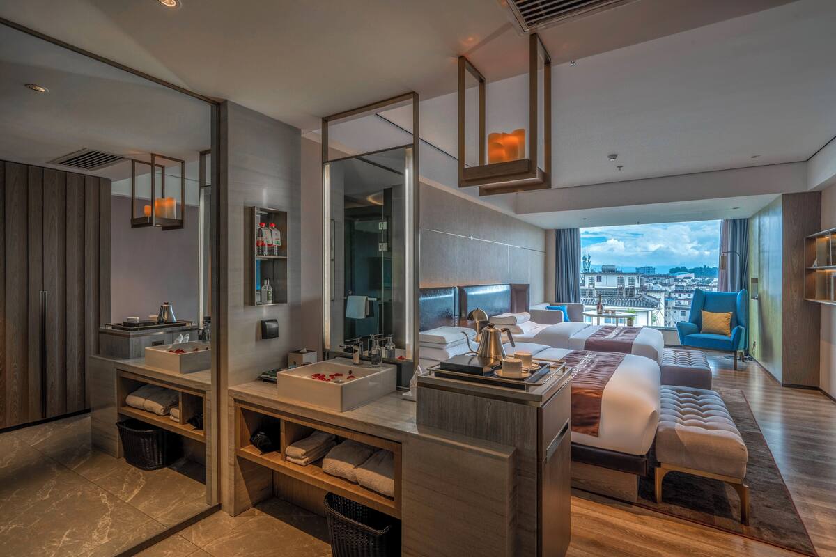 Una habitación de hotel moderna con una cama grande, un área de estar separada, un baño con un espejo amplio y vistas a la ciudad.