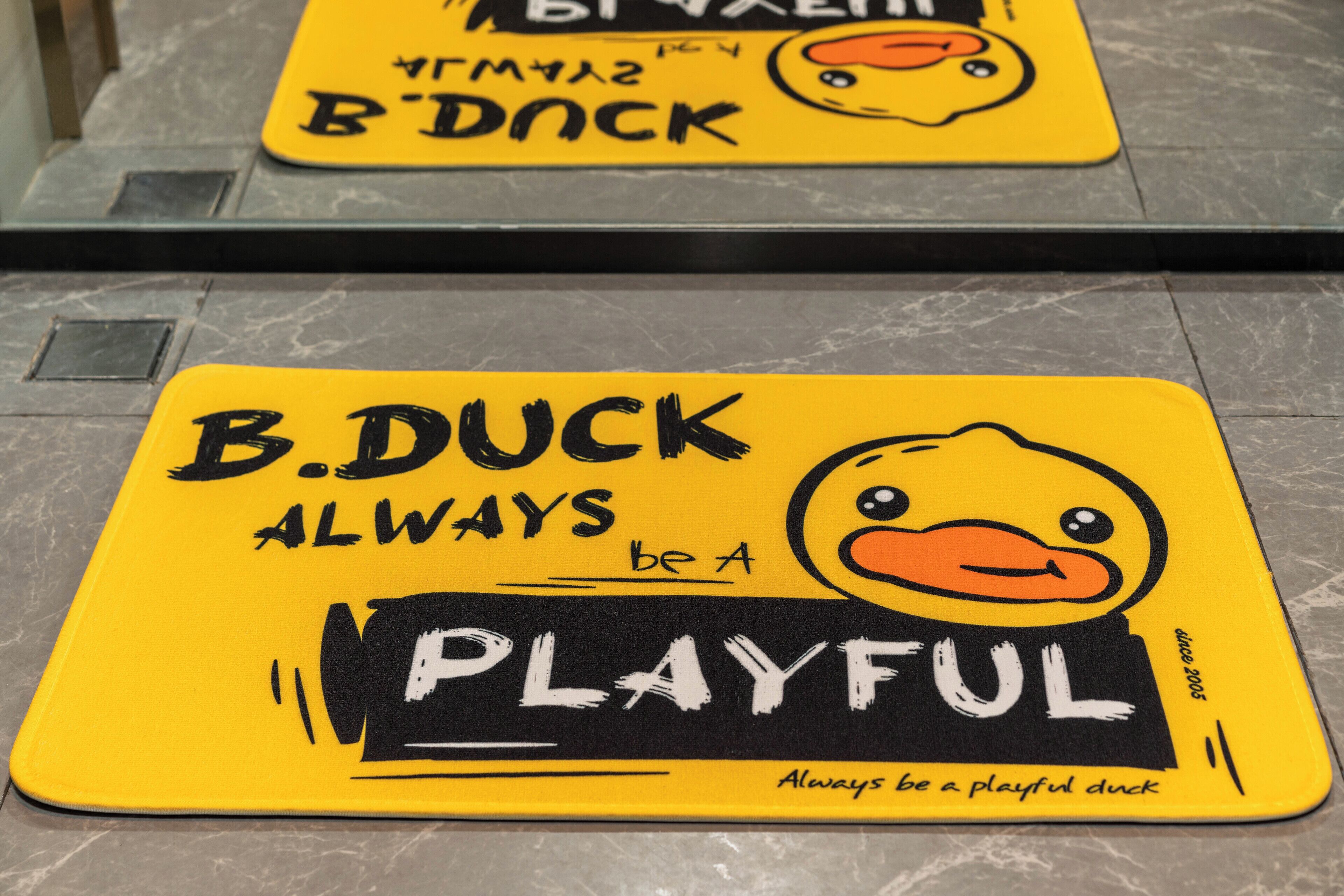 A yellow mat with a duck illustration and the text 'B.DUCK ALWAYS be a PLAYFUL duck' on a tiled floor.