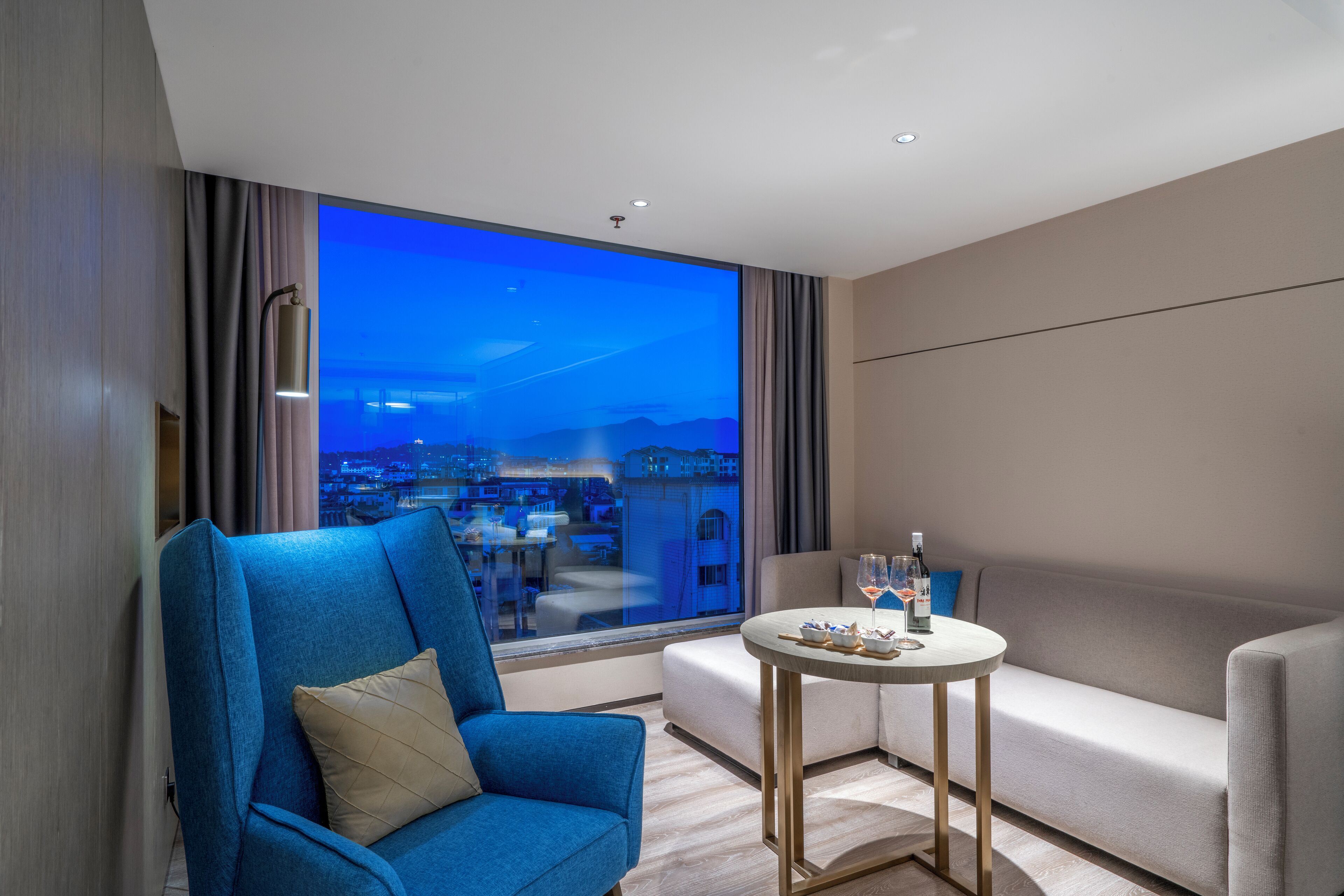 A modern hotel room with a large window offering a city view, a blue armchair, a white sofa, and a small round table with a bottle and glasses.