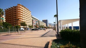 Ein gepflasterter Weg mit einer Reihe mehrstöckiger Gebäude, Palmen und einem überdachten Bereich mit Bänken.